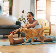 A woman stretches on a yoga mat while an orange cat rubs against her arm. 